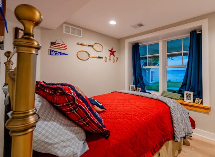 Მხიარული და პატრიოტული საძინებელი ვინტაჟური საჩოგბურთო რეკეტებით, გამუქებული წითელი და ლურჯი თეთრეულითა და მყუდრო ტბის ხედით ფანჯრის კუთხიდან.
