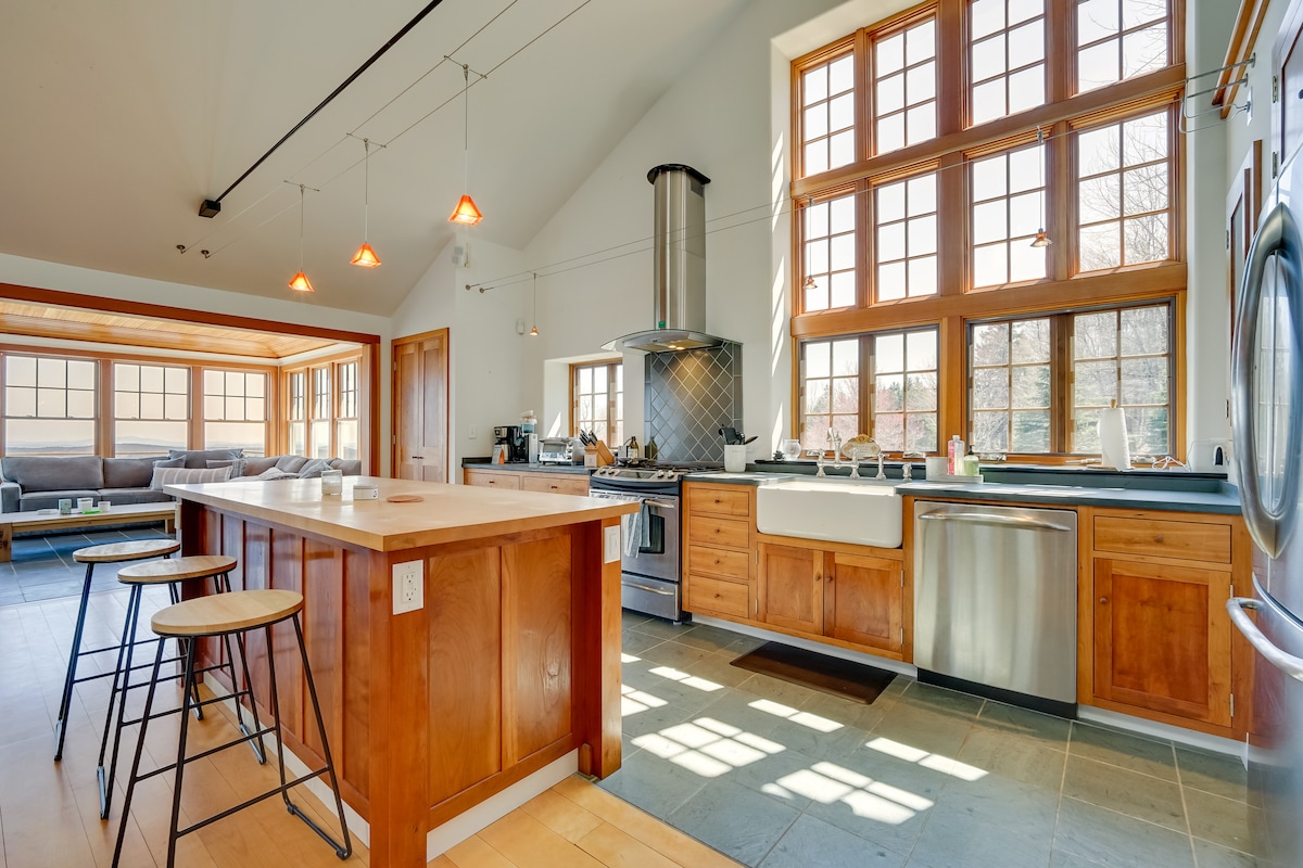 A spacious kitchen features a central island with bar seating, illuminated by pendant lights. Large windows provide natural light and views of the outdoors. The kitchen is equipped with stainless steel appliances, including a gas range and a farmhouse sink, complemented by wooden cabinetry.