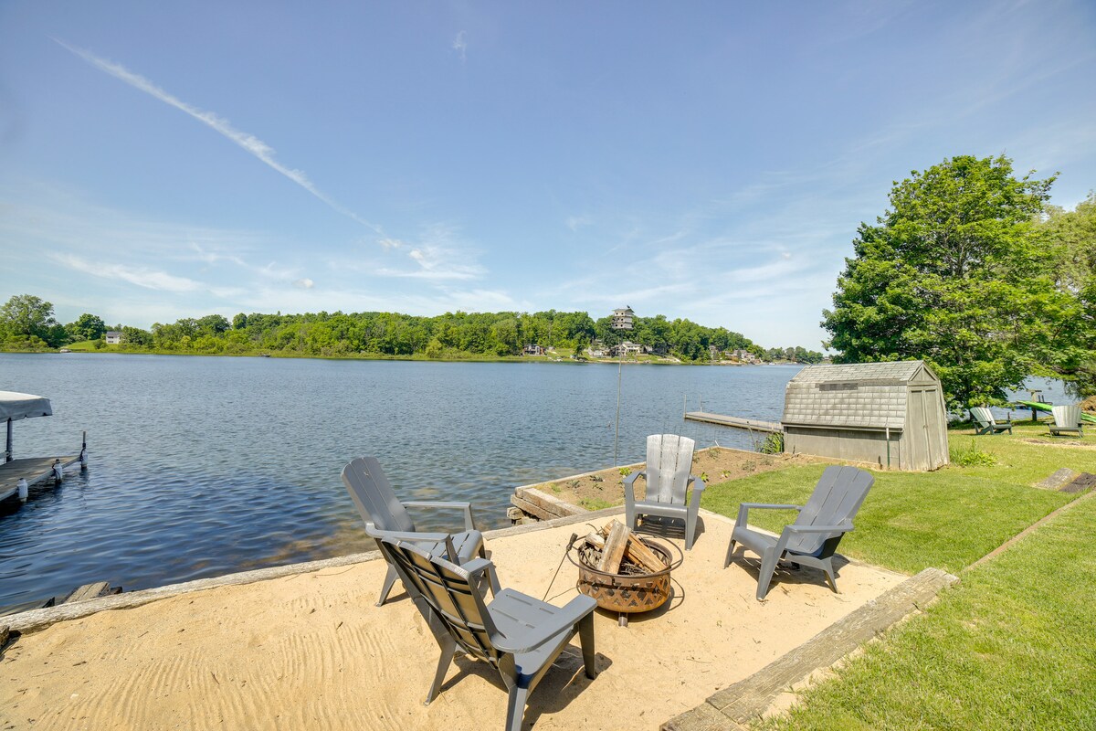 A sandy area by the lake is surrounded by four gray Adirondack chairs arranged around a fire pit. The calm lake is visible in the background, reflecting the clear blue sky and green trees along the shoreline.