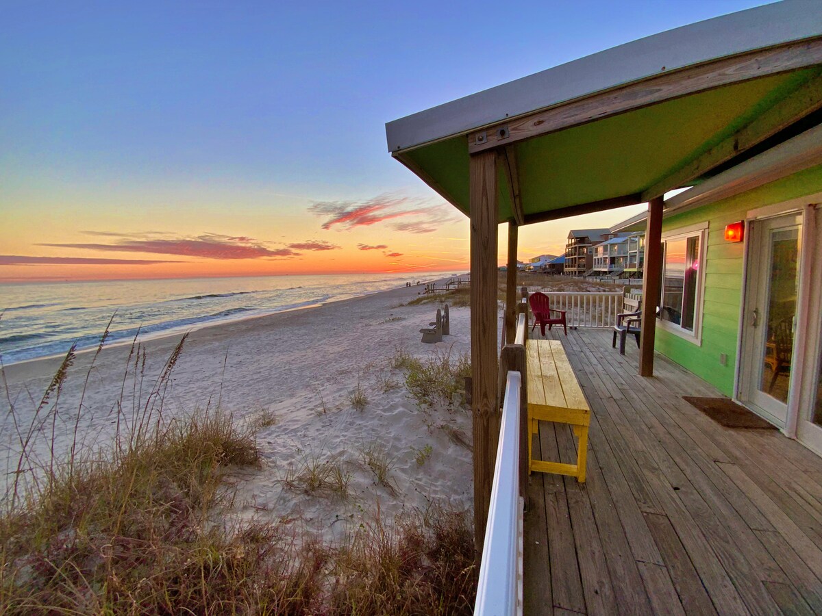 A beachfront deck area showcases a yellow picnic table and Adirondack chairs on wooden flooring. Soft sand and gentle waves are visible nearby, while a pastel sunset paints the sky in hues of pink and orange, contributing to a serene seaside atmosphere.