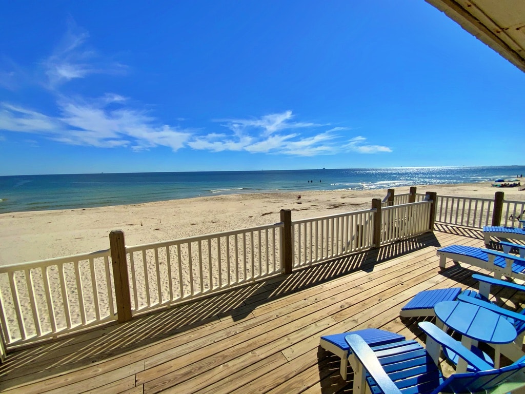 An expansive deck overlooks the sandy beach and calm Gulf waters under a clear blue sky. Several blue lounge chairs are positioned for relaxation, while a white railing provides a safe boundary. The serene landscape includes gentle waves and distant boats on the horizon.