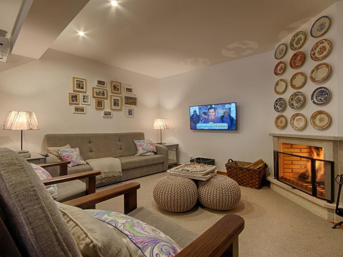 A serene living area features a soft, neutral color palette complemented by cozy furnishings. Two comfortable armchairs and a sofa are positioned around a low table. Decorative plates adorn the wall above a fireplace, which adds warmth to the space, while a TV is mounted nearby.