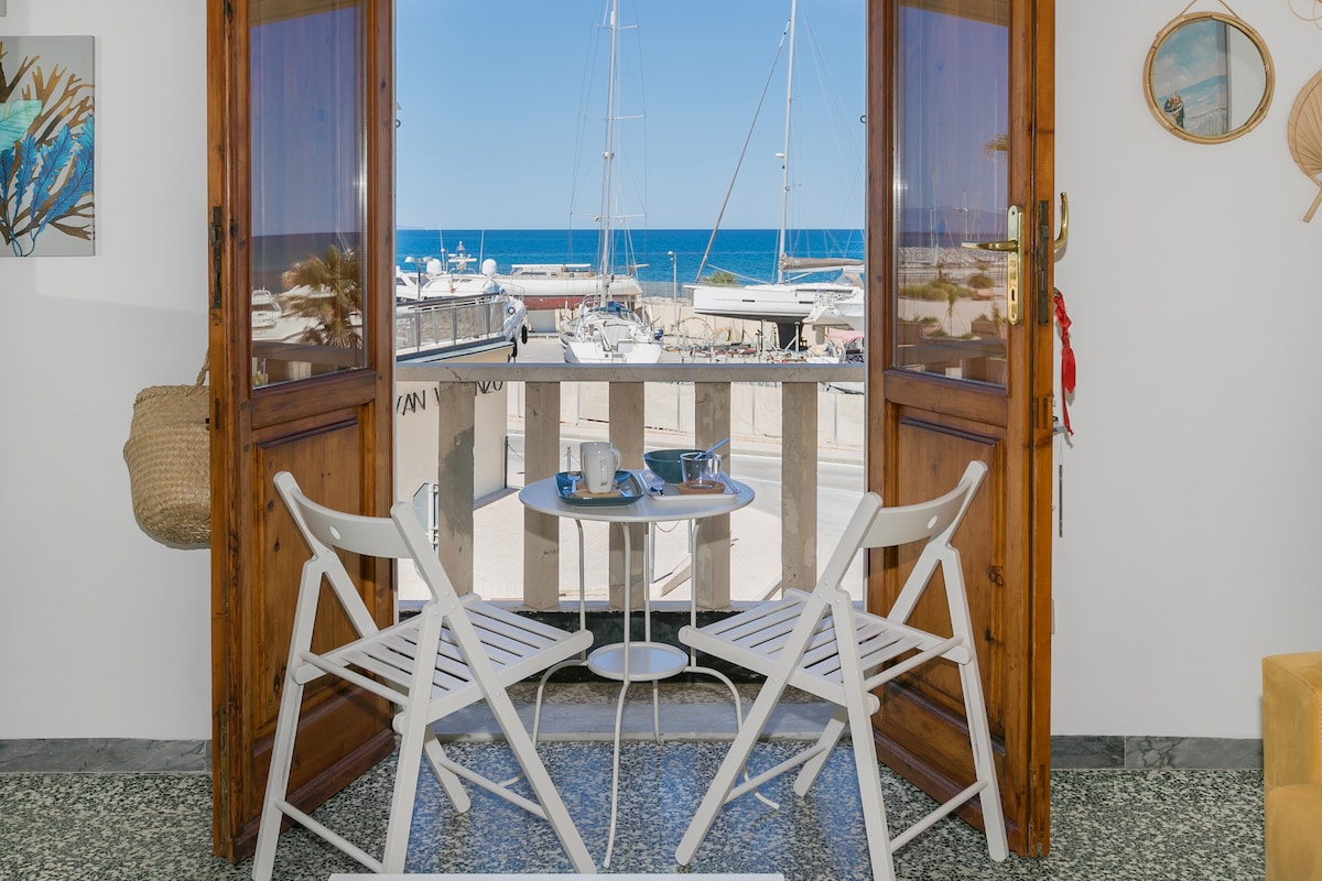 A small balcony features two white folding chairs and a round table, offering views of the marina and the sea. Bright blue water and boats are visible beyond the railing, with clear skies enhancing the coastal ambiance.