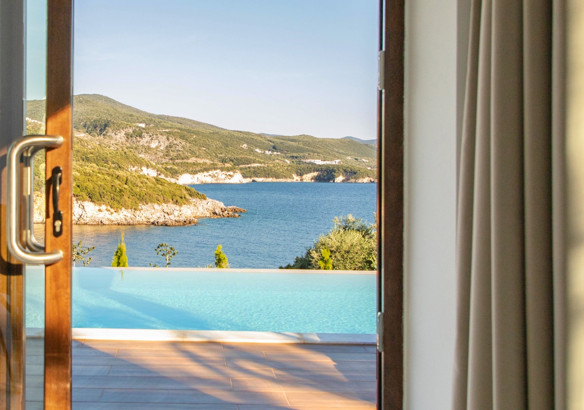 An inviting view from the villa's door frames a serene outdoor area featuring a private infinity pool that blends into the ocean. Lush greenery and the distant hills create a peaceful backdrop under clear blue skies.