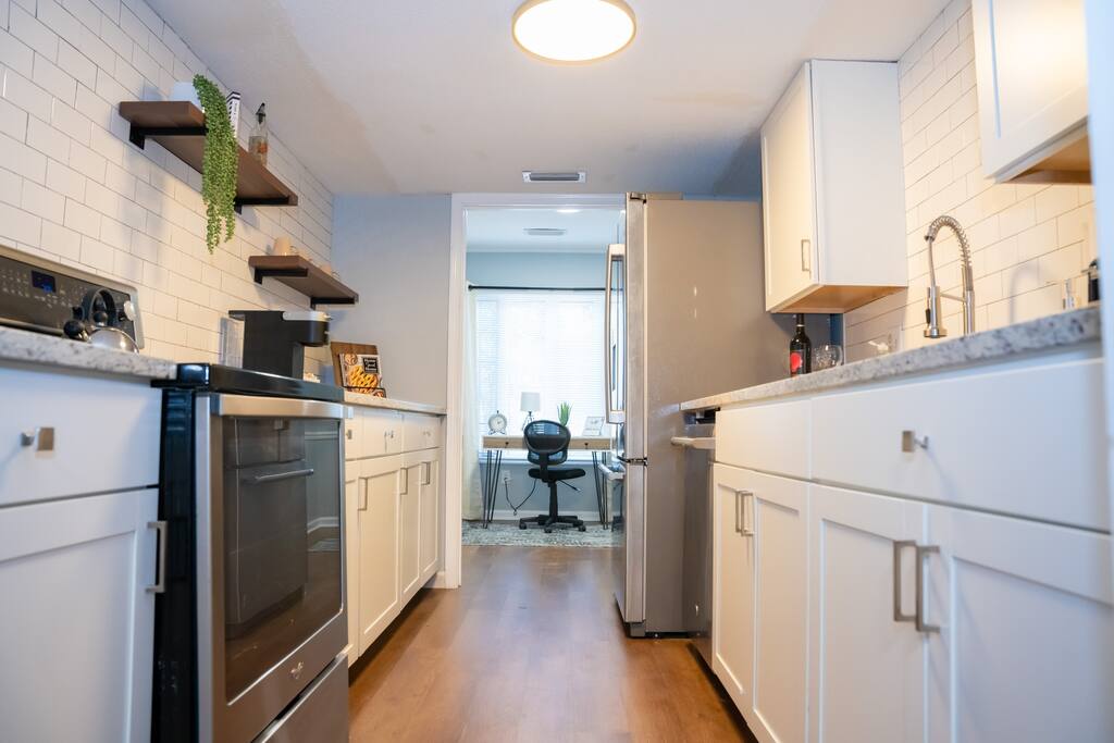 A modern kitchen layout features white cabinetry and a sleek countertop. Stainless steel appliances, including an oven and refrigerator, are visible. The space is illuminated by overhead lighting, and open shelving adds a touch of decor. A glimpse of a multi-functional office area can be seen in the background.