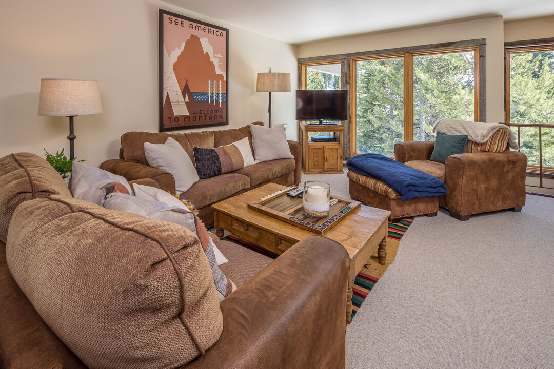 The living area features comfortable sofas arranged around a rustic wooden coffee table. Large windows allow natural light to fill the space, revealing views of the surrounding trees. A flat-screen TV is positioned on a wooden entertainment unit, offering a cozy setting for relaxation.