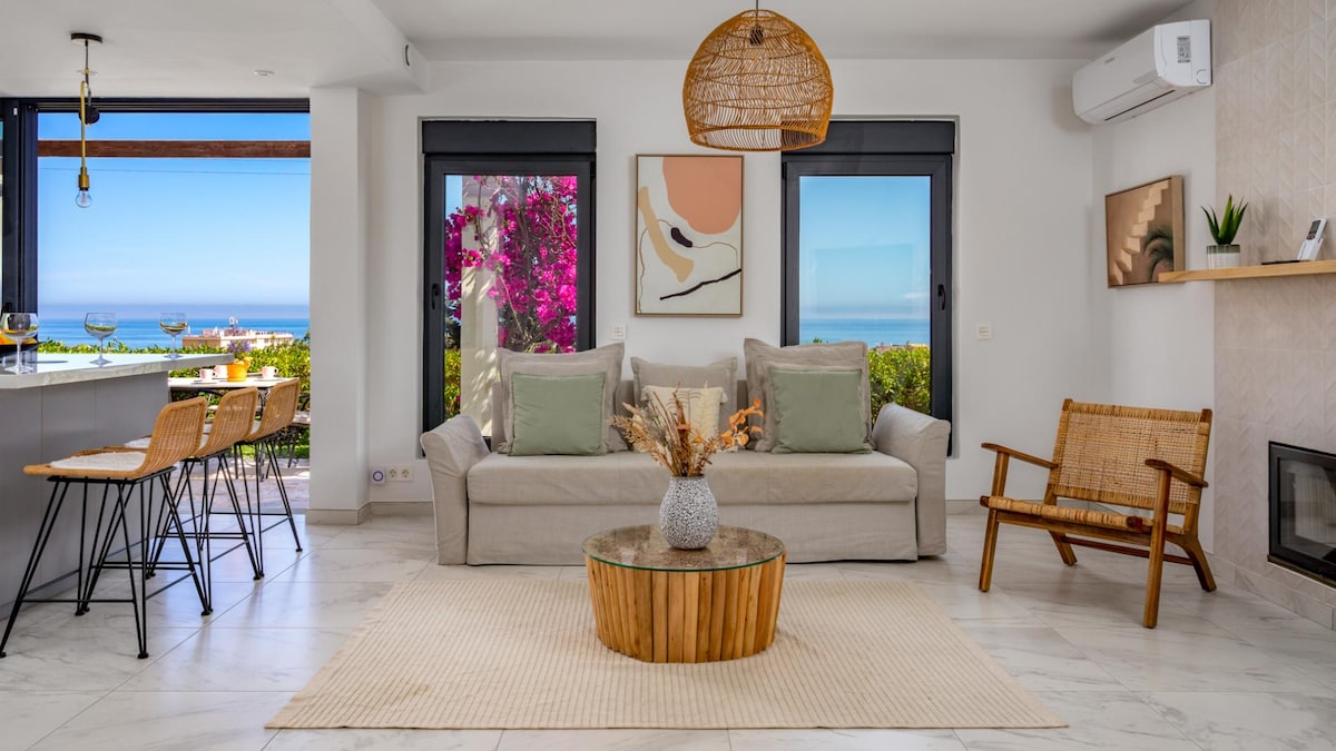 A stylish living area features a comfortable light-colored sofa accompanied by accent cushions. A round wooden coffee table sits centrally on a neutral rug. Large windows offer scenic views of vibrant floral surroundings and distant coastal scenery, while a small dining space includes modern chairs.