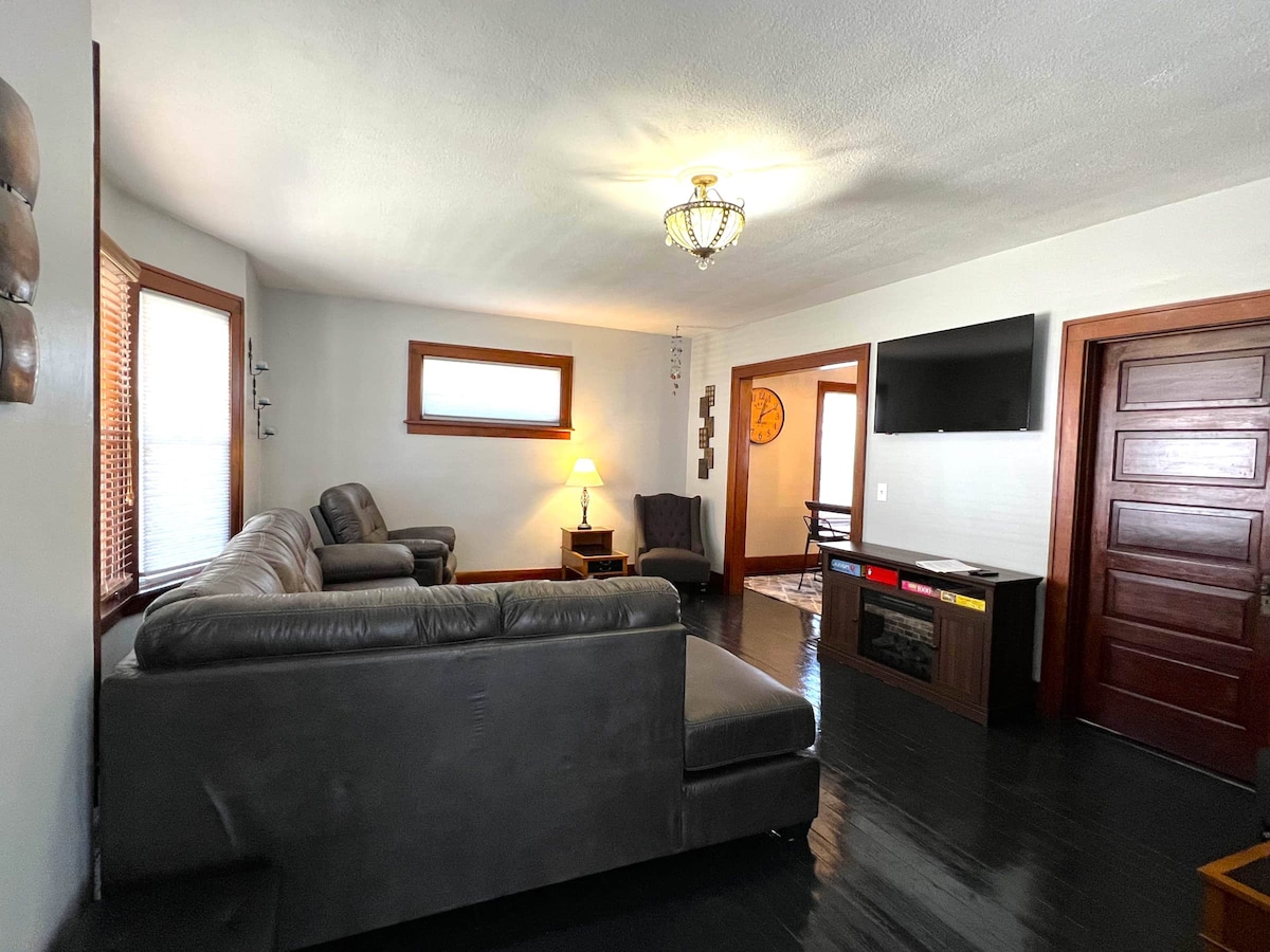 A spacious living room is furnished with a dark sectional sofa and a cozy armchair. Natural light enters through the windows, and a stylish lamp provides additional illumination. A flat-screen TV is mounted on the wall above a modern media console, complementing the warm decor.