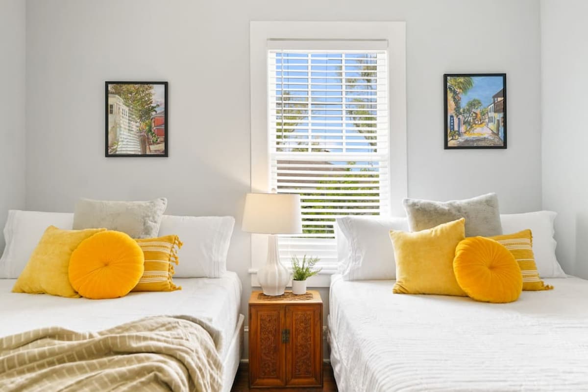 A bright bedroom features two spacious beds topped with white linens and vibrant yellow pillows. A decorative lamp stands on a bedside table, accompanied by a small green plant. Natural light floods the room through a window adorned with white blinds, framed by artwork depicting scenic views.
