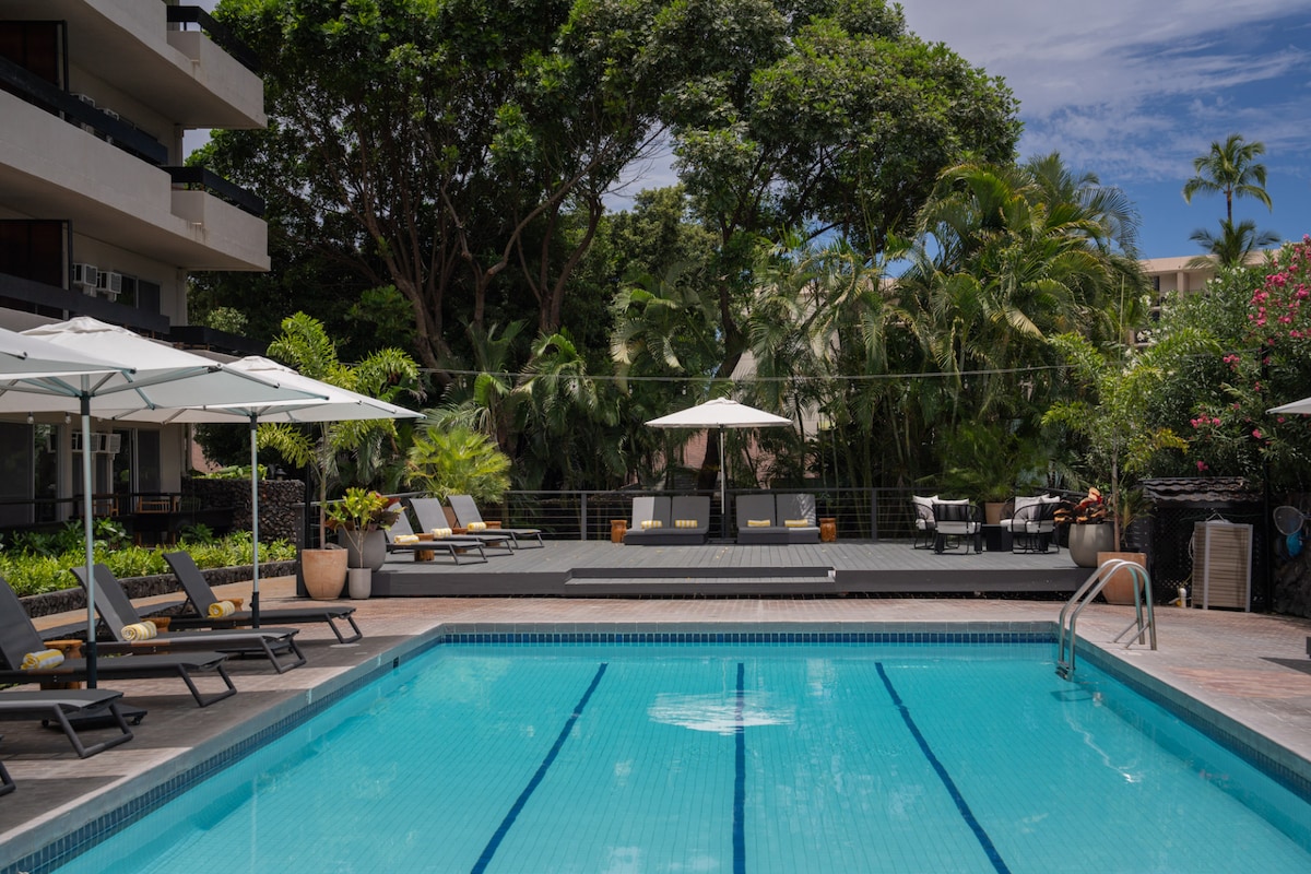 An outdoor swimming pool features clear water surrounded by lounge chairs and umbrellas. Lush greenery and palm trees provide a natural backdrop, while shaded seating areas can be seen in the distance. Inviting spaces create an atmosphere conducive to relaxation and leisure.