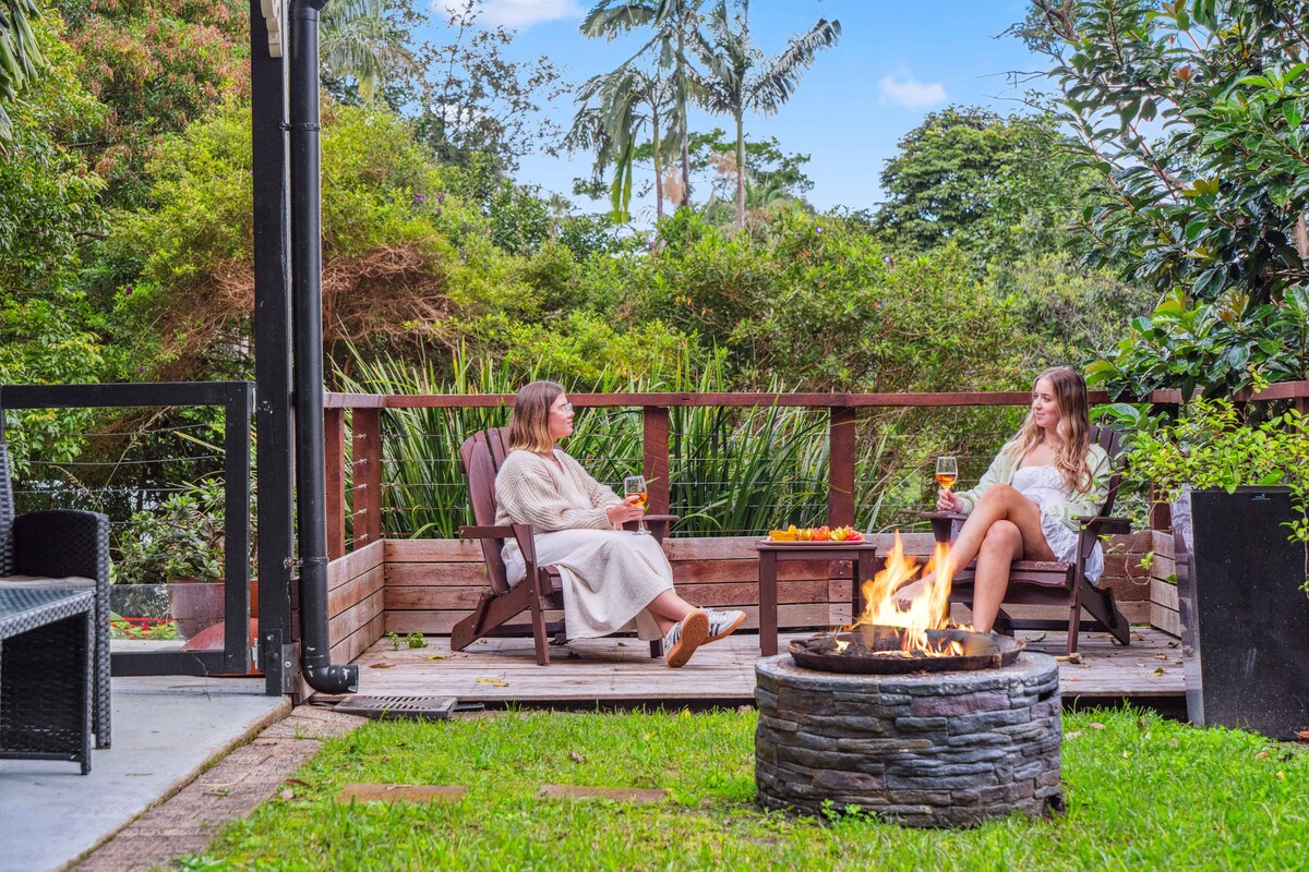 An outdoor deck is depicted, furnished with two adirondack chairs positioned around a fire pit. Surrounded by lush vegetation, the space invites relaxation, with a small table showcasing refreshments. The fire pit is lit, creating a warm ambiance against the verdant backdrop.