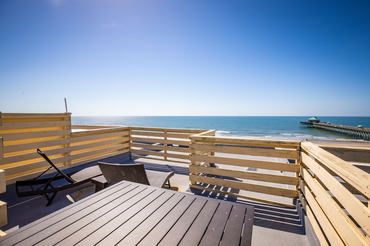 A rooftop deck offers panoramic views of the ocean, with gentle waves lapping against the shore. Wooden railings surround a dining table, and a lounge chair is positioned for relaxation, creating an inviting space to enjoy the warm sun and sea breeze.