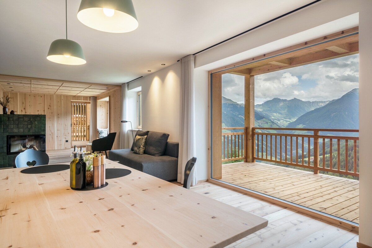 A bright living area features a wooden dining table with glass bottles and a view of the panoramic terrace. The room is enriched with warm wooden textures and a cozy seating space, while large sliding doors open to reveal mountain scenery and a wooden railing.