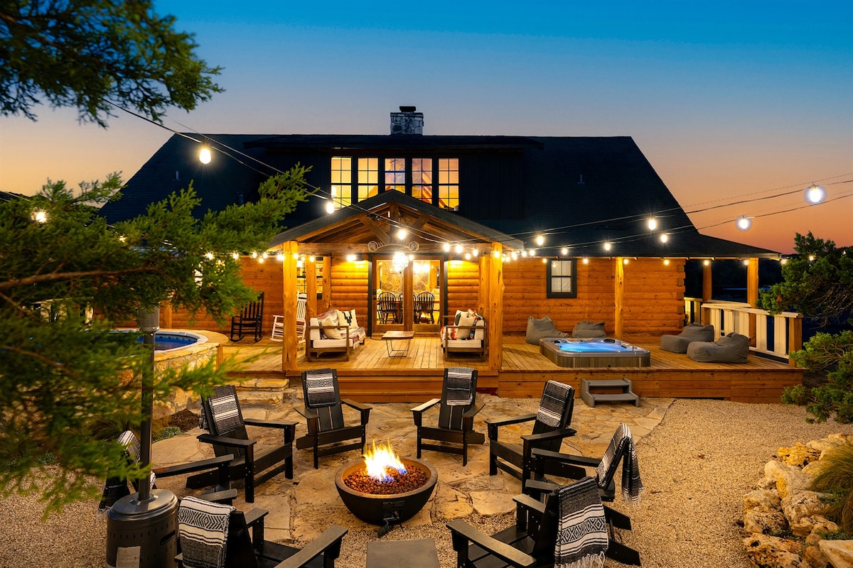 An inviting cabin exterior features warm wooden tones, surrounded by a serene landscape. A circular seating arrangement with black chairs encircles a central fire pit, while string lights create a cozy ambiance. The hot tub and lounge areas are positioned on the spacious deck for relaxation.