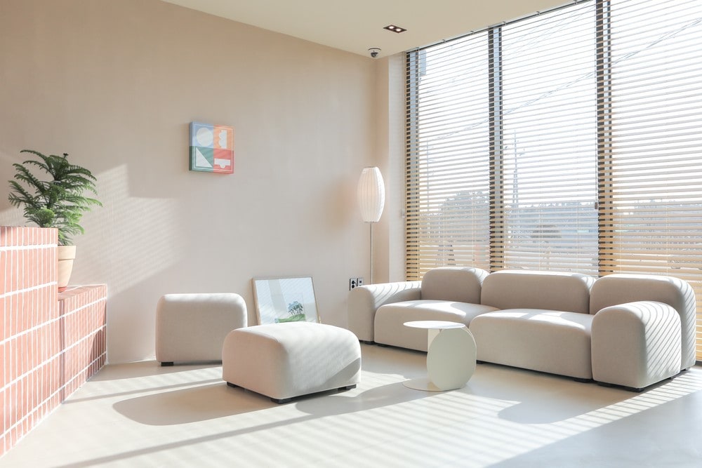 A bright and airy living area is featured, highlighted by large windows adorned with horizontal blinds. A modern, light-colored sofa accompanies a plush ottoman and a minimalist side table. A potted plant adds a touch of greenery beside an art piece mounted on the wall.