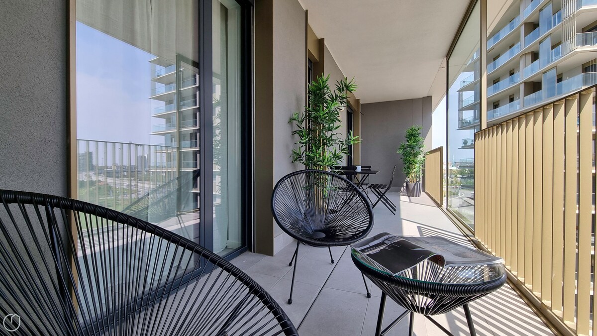 An inviting balcony area features two black chairs with circular designs and a small table. Potted plants add greenery to the space, while sunlight casts shadows across the floor. The railing offers views of the surrounding urban landscape.
