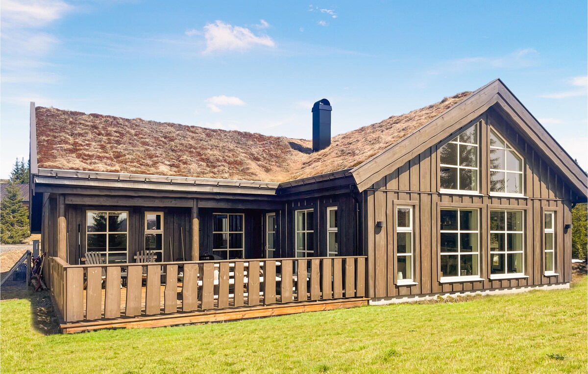 A spacious wooden cabin features a sloped grass roof and a large deck. Floor-to-ceiling windows provide abundant natural light, highlighting the traditional craftsmanship. The surrounding grassy area adds to the serene atmosphere of the mountain setting.
