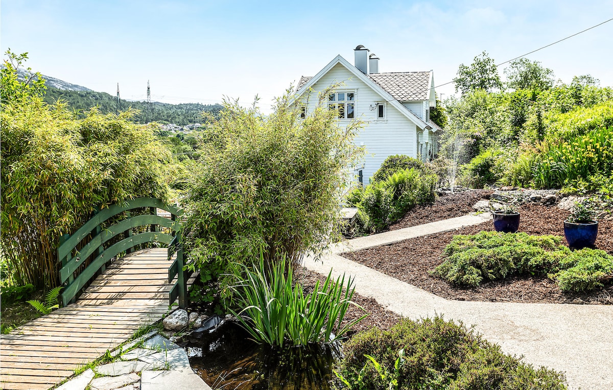 A serene pathway leads to a charming cottage, surrounded by lush greenery and colorful plants. A small wooden bridge extends over a tranquil water feature, enhancing the peaceful atmosphere. The landscape is bathed in natural light, showcasing the inviting outdoor space.