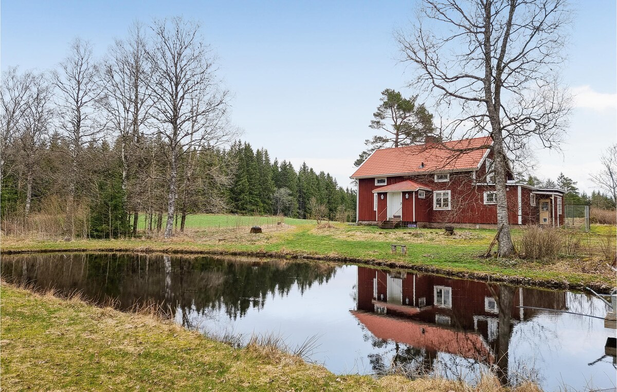A red vacation home is reflected in a serene pond, surrounded by lush greenery and trees. The rustic structure features a sloped roof and multiple windows, set against a backdrop of open fields and woodland. Calm waters create a tranquil atmosphere.