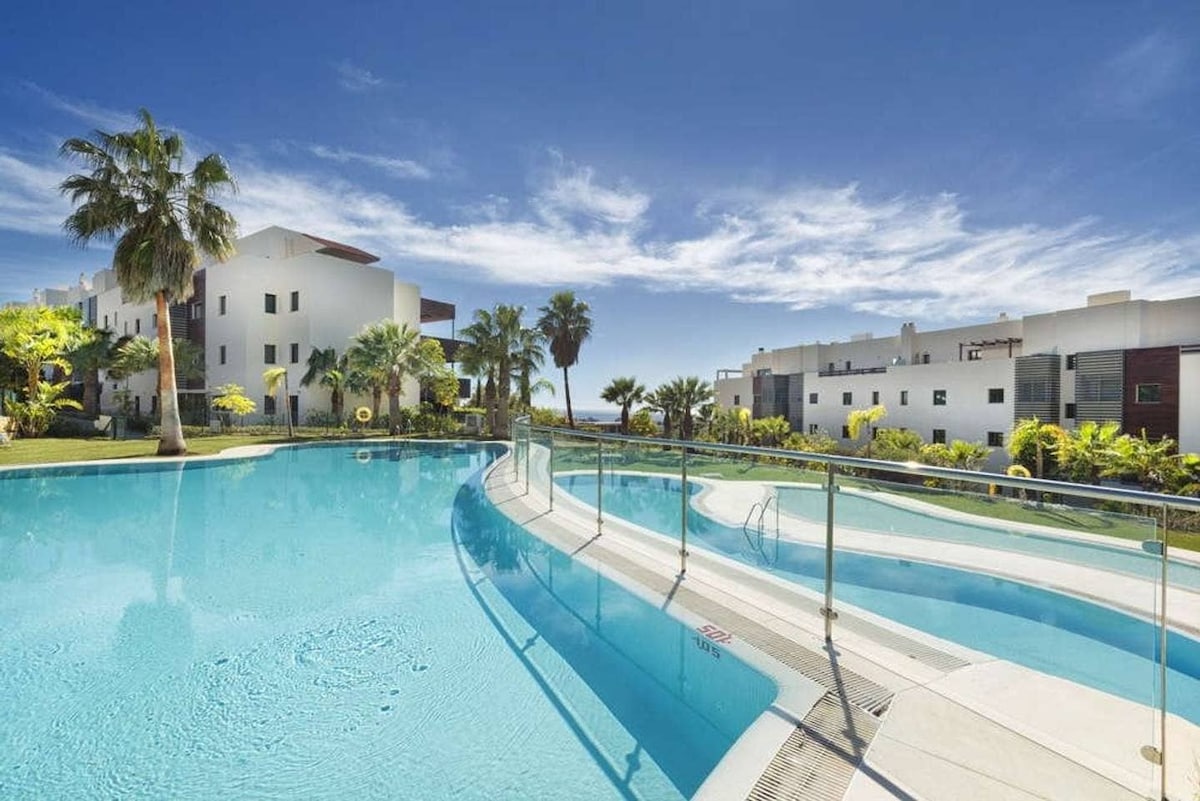 A spacious lagoon-style swimming pool is featured, with gentle curves and clear blue water inviting relaxation. Lush palm trees and mature landscaping surround the area, providing a tranquil environment. Bright blue skies complement the serene atmosphere, enhancing the view of the surrounding residential buildings.