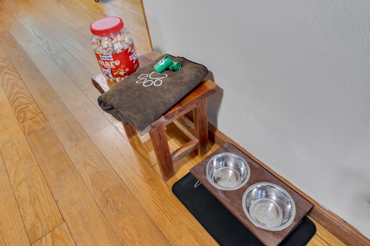 A small wooden stool is positioned next to a dark mat, holding a container of treats for pets and a green toy. Two stainless steel bowls are placed on a wooden base, providing a designated area for feeding pets.