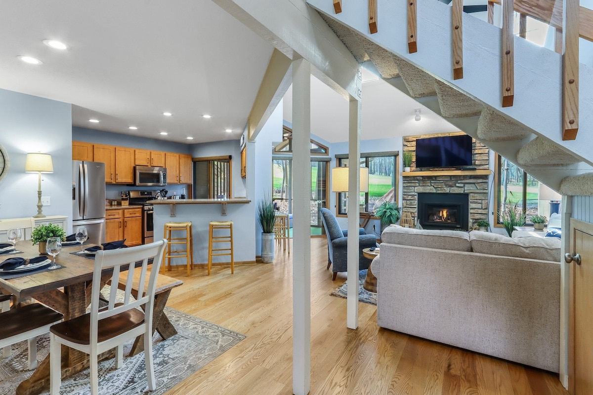 An open-concept living area features a dining table set for four and a cozy seating arrangement with a fireplace nearby. Natural light floods the space through large windows, and a staircase leads to the upper level while lush greenery is visible outdoors.