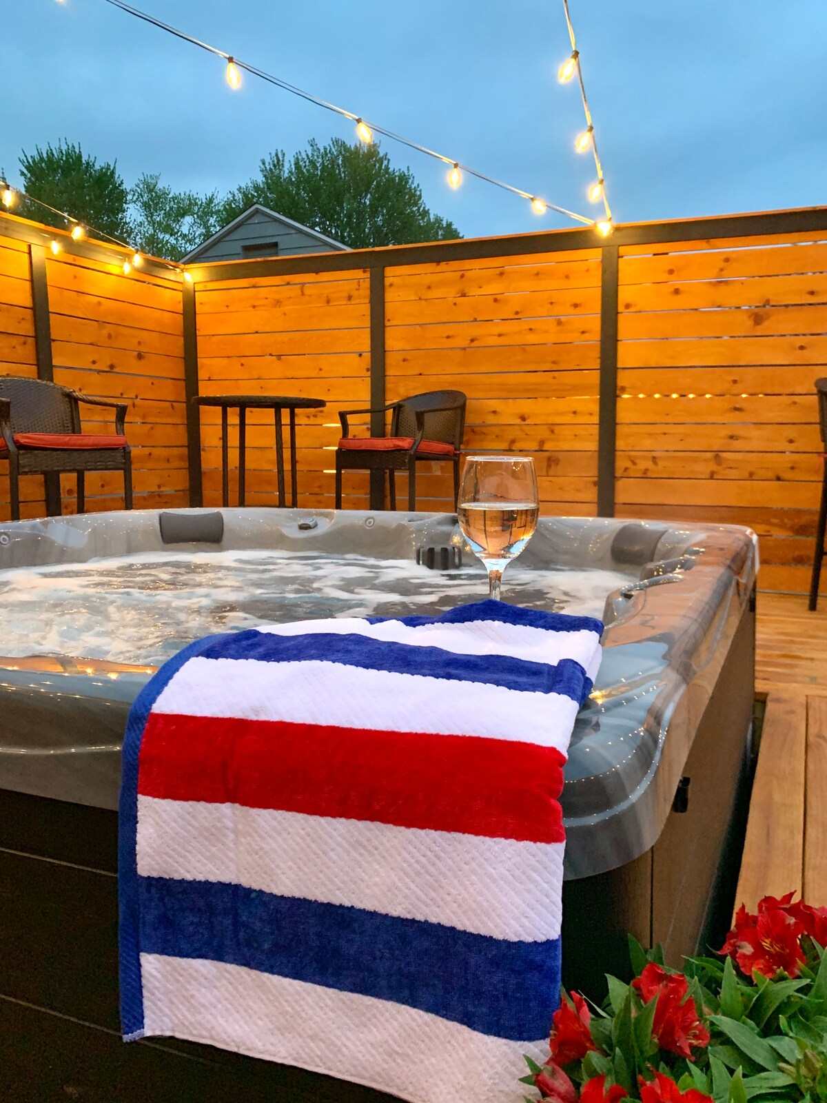 A hot tub is filled with bubbling water, accompanied by a towel draped over the edge in red, white, and blue stripes. Soft lighting from string lights above creates a warm ambiance in the enclosed deck space, featuring outdoor seating for relaxation.