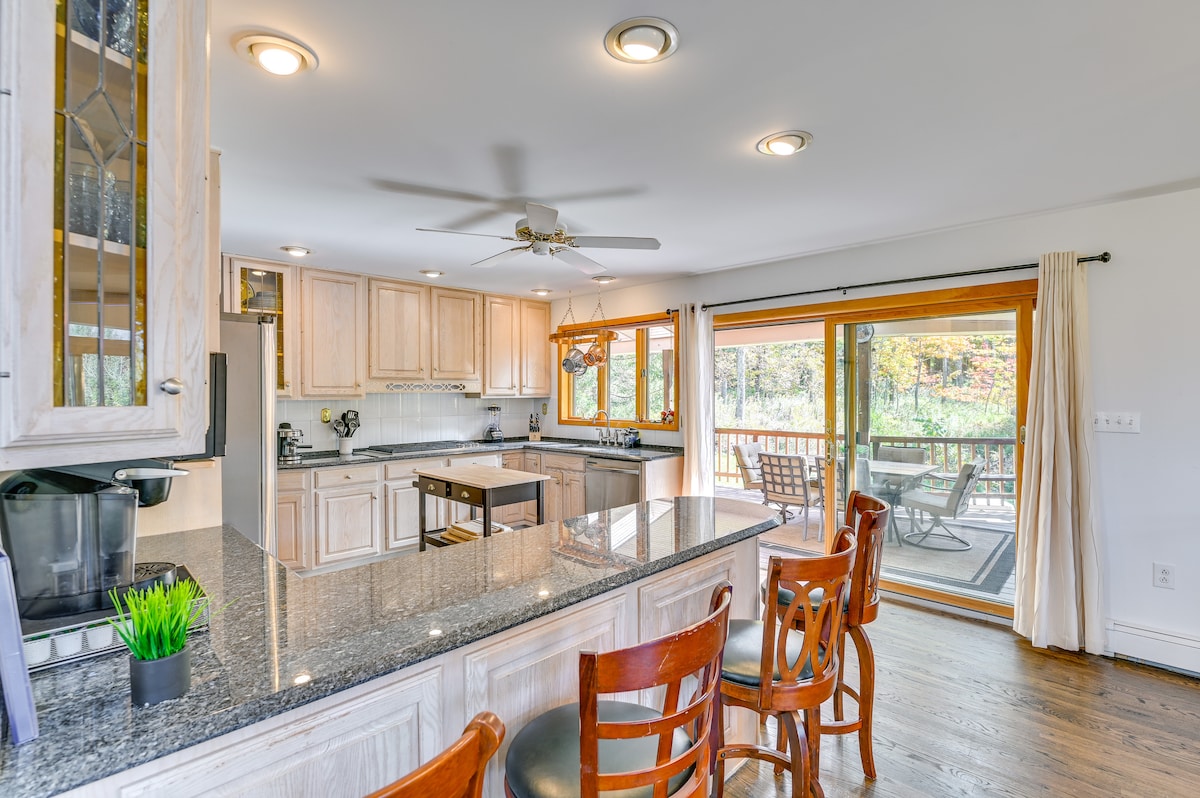 A light and airy kitchen area features hardwood flooring and a central island with seating for four. Wooden cabinetry provides ample storage, and large sliding doors open to a deck overlooking the trees. Natural light floods the space, enhancing its inviting atmosphere.
