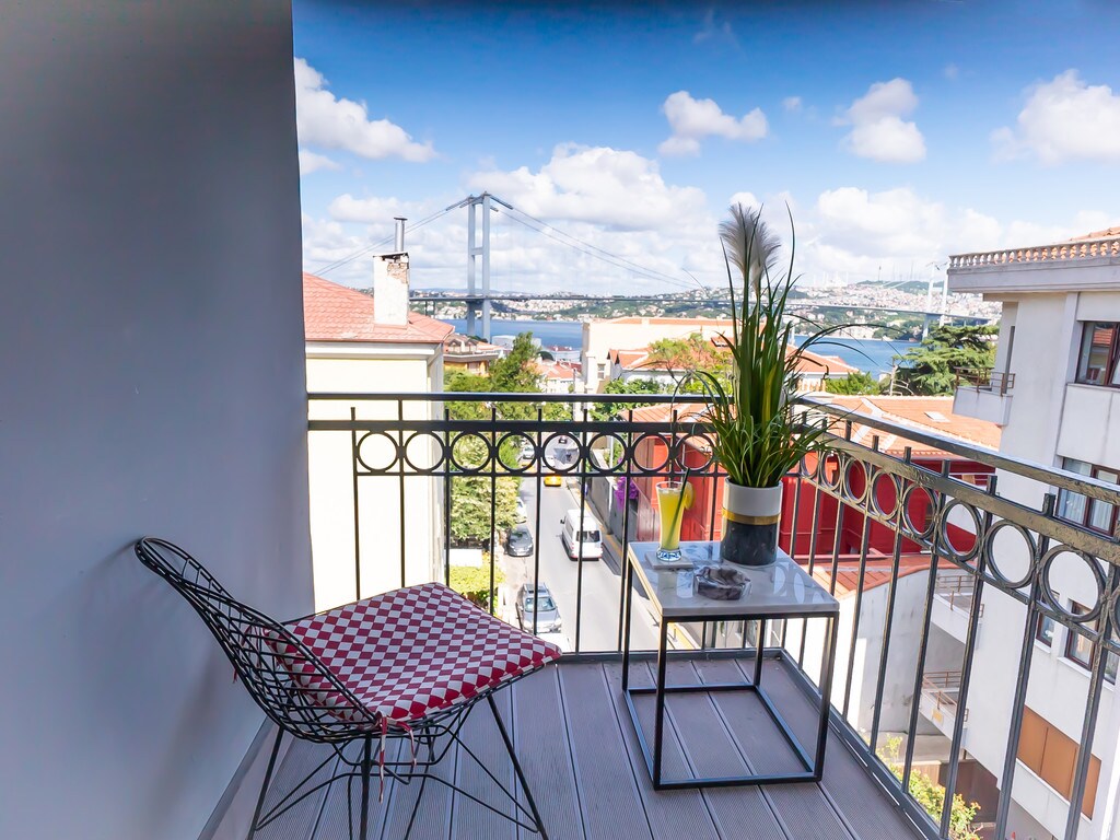 A small balcony is presented with a wire chair and a table featuring a decorative plant. The vibrant view includes the Bosphorus Bridge and the surrounding rooftops, with expansive sky and scattered clouds enhancing the atmosphere.