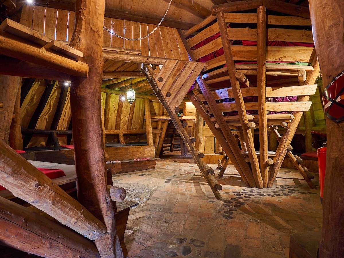 The interior features a unique design with wooden beams and rustic furnishings. A staircase made of logs leads to upper sleeping areas, while stone flooring adds texture. Soft red bedding is visible on elevated sleeping platforms, providing an inviting space for rest.