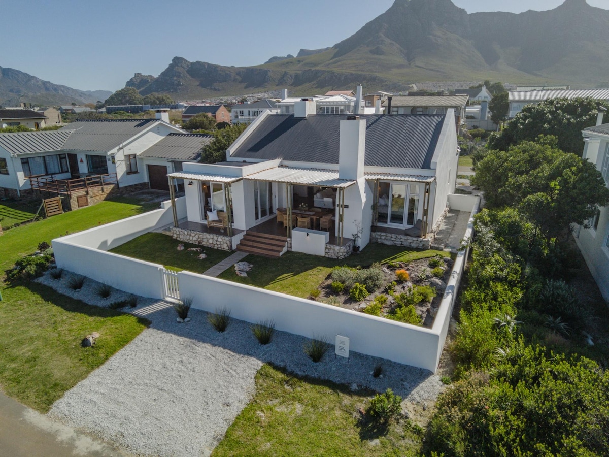 A modern cottage is showcased with a light-colored exterior and black roof, surrounded by a landscaped garden. The patio is partially covered, providing shaded outdoor space. Scenic mountains rise in the background, with a clear blue sky overhead and a combination of gravel and greenery in the foreground.