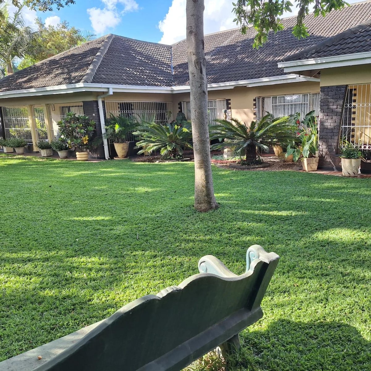 A view of the guest house showcases a well-maintained green lawn, bordered by various potted plants. A bench is positioned in the foreground, offering a spot to relax and enjoy the serene garden atmosphere beneath a blue sky scattered with clouds.