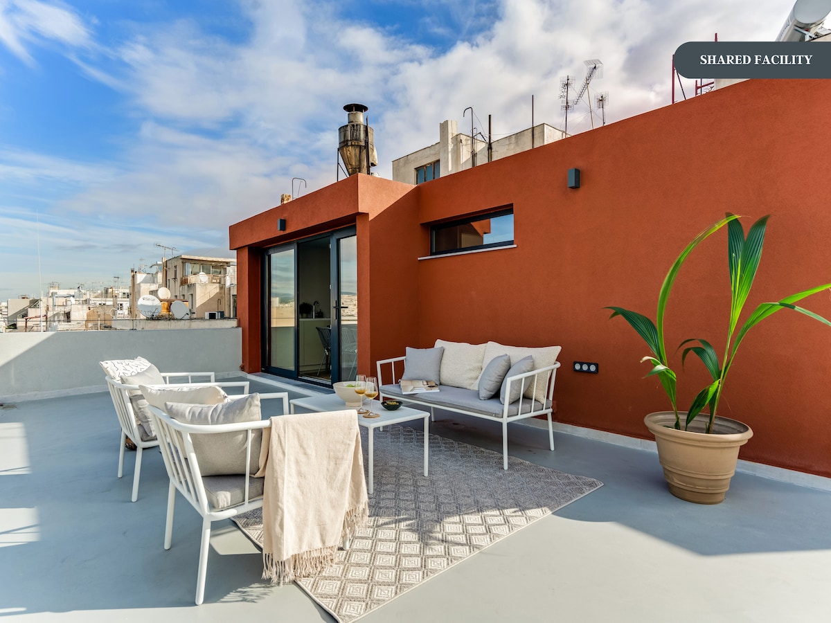 A spacious rooftop terrace is adorned with a cozy seating area that includes a light-colored sofa and four white chairs. A large potted plant adds a touch of greenery, while an intricate rug defines the seating space. The skyline of the city is visible in the background under a clear sky.