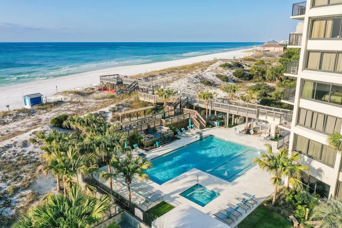 An aerial view captures the expansive saltwater pool bordered by palm trees and lounging chairs. The pool area is surrounded by a well-maintained deck leading to a private walkway that provides direct access to the white sandy beach and the emerald waters of the Gulf.