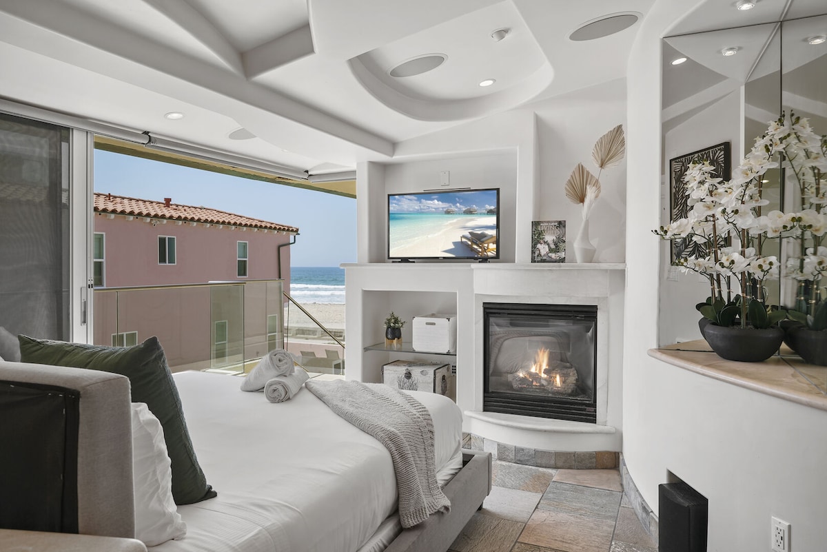 A cozy sleeping area features a spacious bed with soft linens, complemented by a modern fireplace. A flatscreen television is mounted above the fireplace, while large glass doors frame an unobstructed view of the ocean. Decorative elements include potted plants and elegant wall accents.