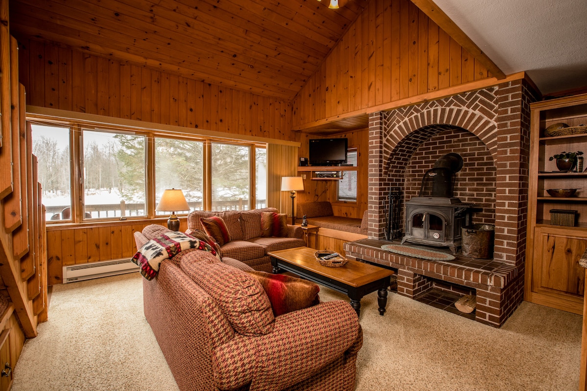 A warm living area is highlighted by rich wood paneling and large windows offering natural light and views of the outdoors. A cozy fireplace is set in a brick alcove. Comfortable seating is arranged around a central coffee table, creating an inviting space for relaxation.