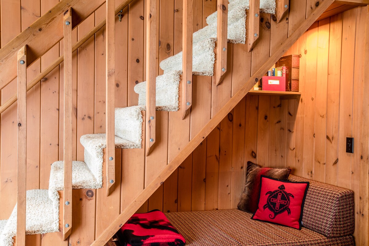 A staircase with wooden railings leads to the upper level, featuring carpeted steps for comfort. A cozy reading nook is placed nearby, adorned with a decorative pillow. Warm wooden panels create a soothing backdrop for the inviting space.