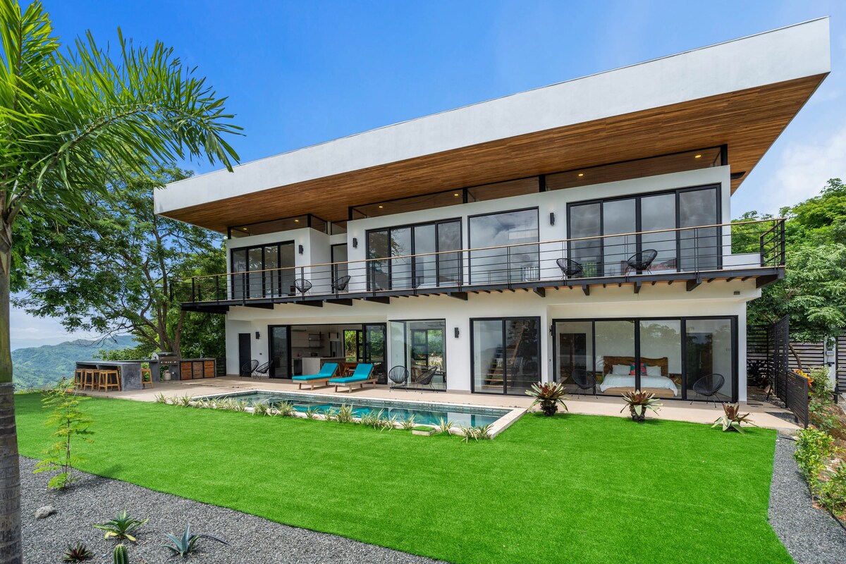 The modern home presents a sleek exterior with two levels, large windows, and a spacious terrace. A tranquil pool is framed by a well-manicured lawn featuring tropical landscaping. An outdoor dining area and seating are visible, inviting relaxation and leisure in the fresh air.