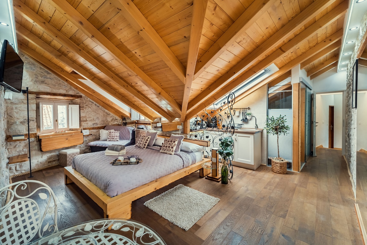 The spacious sleeping area showcases a large wooden bed framed by sloped ceilings with exposed beams. Natural light filters through skylights, illuminating the wooden floor. A cozy seating area includes decorative cushions, while an indoor plant adds a touch of greenery. The room's design combines texture and simplicity.