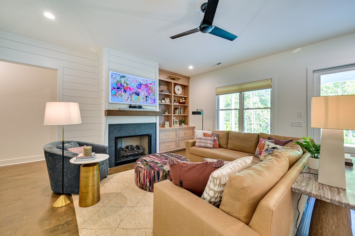 An inviting living area features a comfortable sectional sofa in neutral tones, accented by colorful throw pillows. A round pouf sits in the center, while a contemporary lamp and a wooden side table add a modern touch. A wall-mounted TV is positioned above a gas fireplace, complemented by large windows that provide natural light.