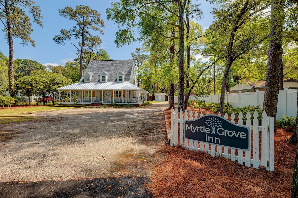 The exterior of the house is framed by tall trees and a manicured yard, revealing a charming building with a gabled roof and a welcoming front porch. A white picket fence surrounds the property, displaying a sign that reads 'Myrtle Grove Inn.'