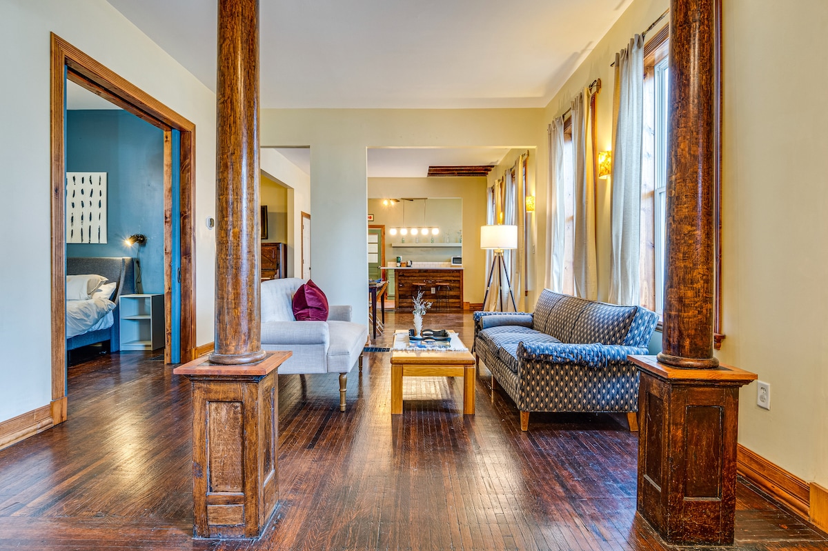 An open living space features polished wooden floors and tall columns that define the area. Cozy seating options include a patterned sofa and a light-colored couch, complemented by a coffee table. Natural light filters through large windows, enhancing the inviting atmosphere.