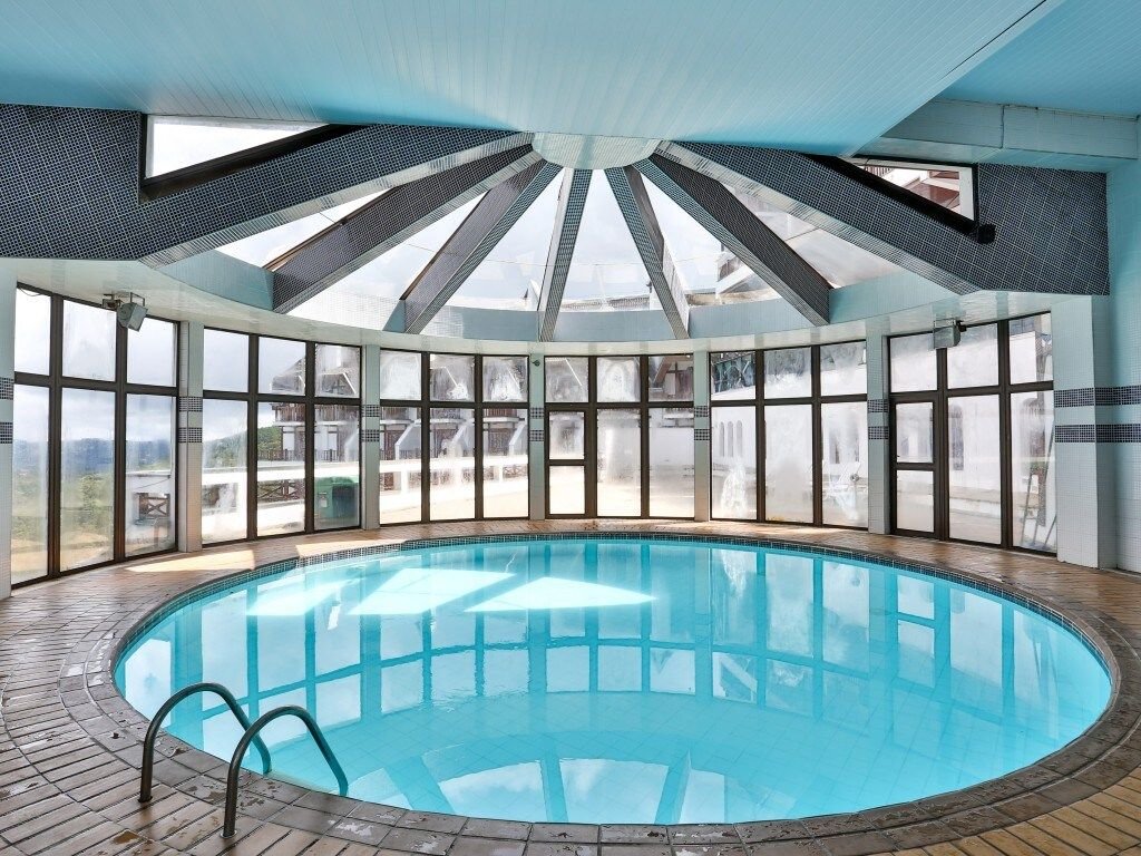 An indoor pool is framed by expansive glass windows, allowing natural light to fill the space. The circular pool features a smooth surface, and a subtle blue hue is seen in the water. A star-shaped skylight enhances the modern design of the area.