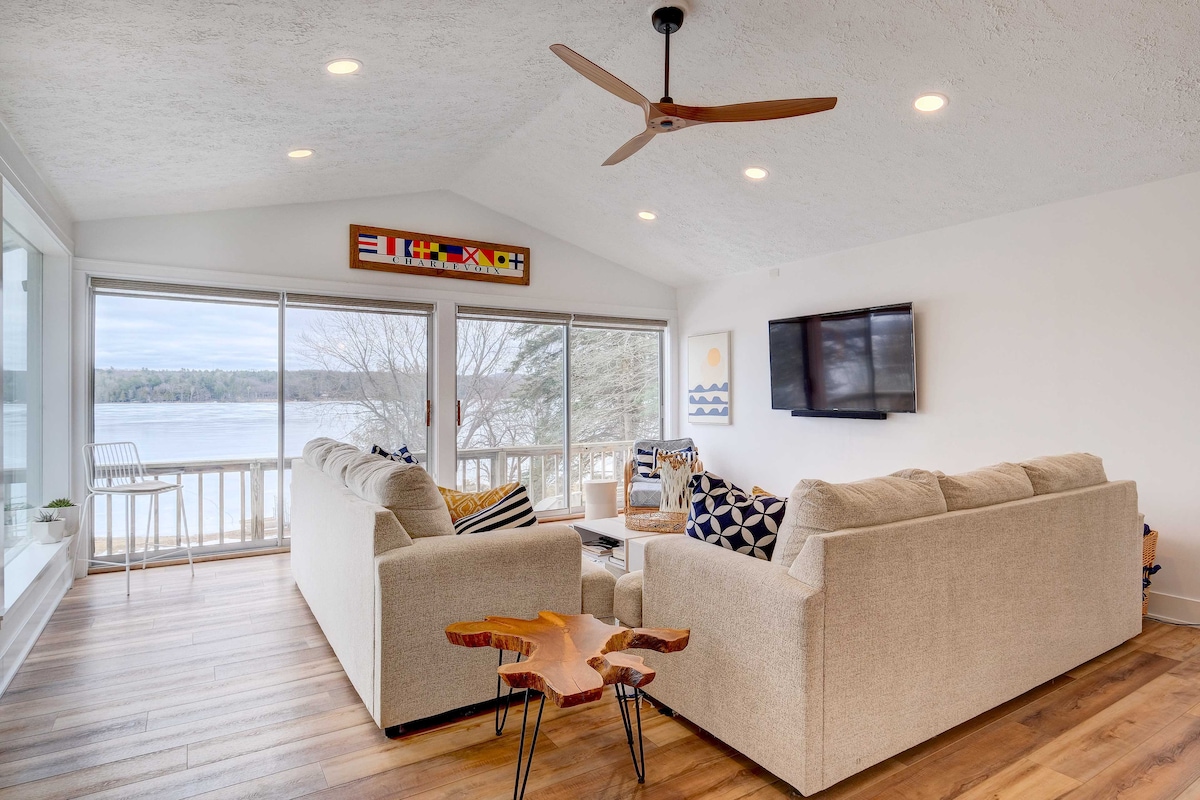 A spacious living area features comfortable seating arranged around a central coffee table. Large sliding glass doors provide a scenic view of the lake, and natural light fills the room. A ceiling fan and a wall-mounted TV add to the modern amenities.