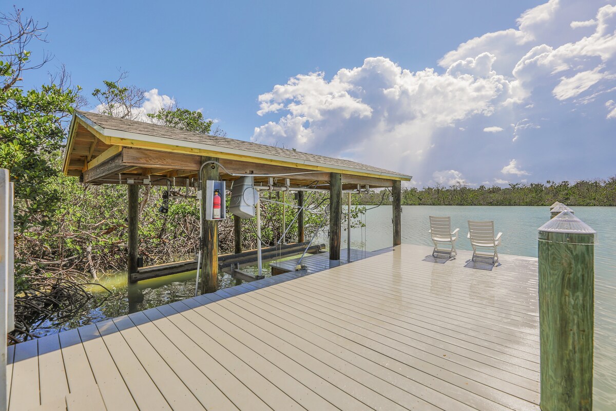 A private dock extends over the tranquil water, surrounded by lush greenery. Two white chairs are positioned on the deck, inviting relaxation while enjoying the scenic views. Sunlight reflects off the surface, creating a serene setting for water activities or peaceful contemplation.