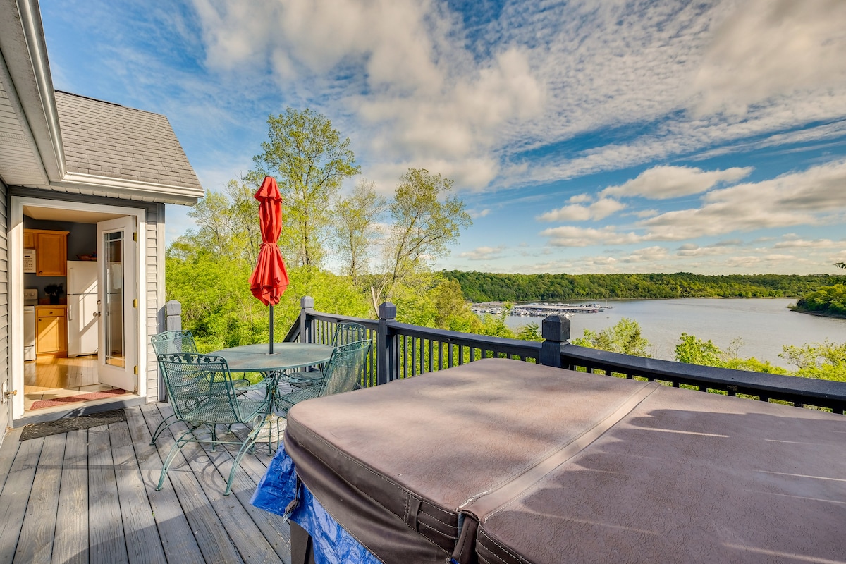 A spacious outdoor deck features a private hot tub and a dining area with green and red accents. Scenic views of the lake and surrounding greenery are visible, highlighted by a partially cloudy sky. Easy access to the indoor kitchen can be observed in the background.