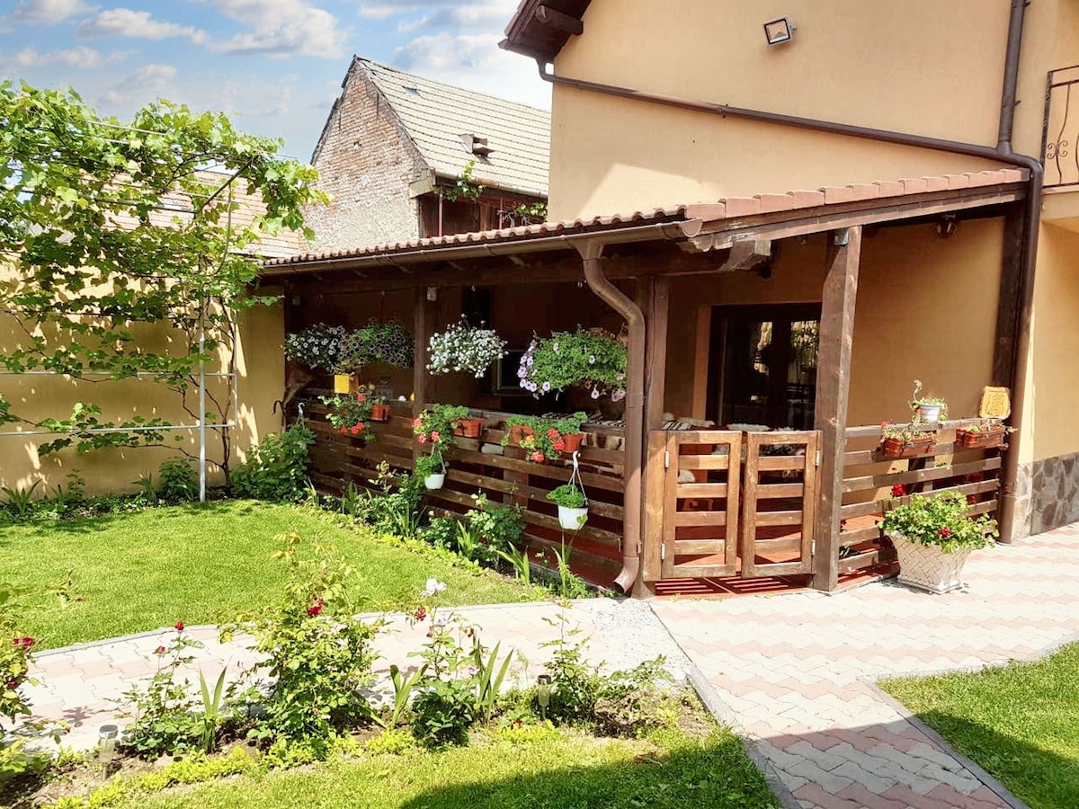 The exterior features a charming terrace surrounded by a wooden railing, adorned with hanging flower pots. Lush greenery in the garden offers a serene atmosphere, complemented by a well-maintained pathway leading to the entrance of the house.