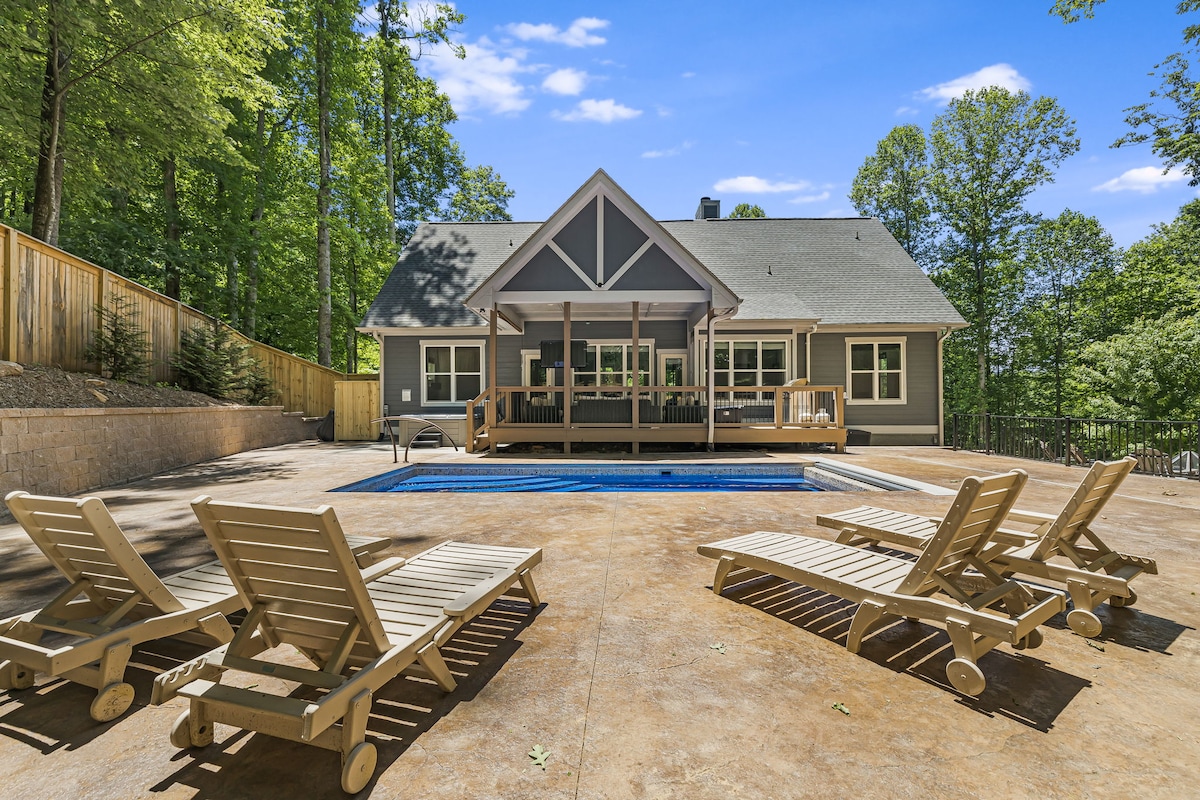 A serene outdoor space features an in-ground pool surrounded by lounge chairs, set against a backdrop of lush greenery. The modern home with a gabled roof is visible behind the pool, offering a spacious deck area for relaxation and socializing.