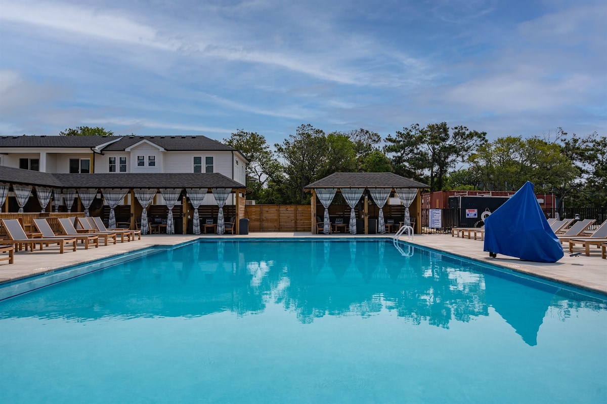 A large heated salt-water pool is surrounded by comfortable lounge chairs and shaded cabanas. The clear water reflects the blue sky overhead, creating a serene atmosphere. A covered area with seating is visible in the background, providing additional options for relaxation.