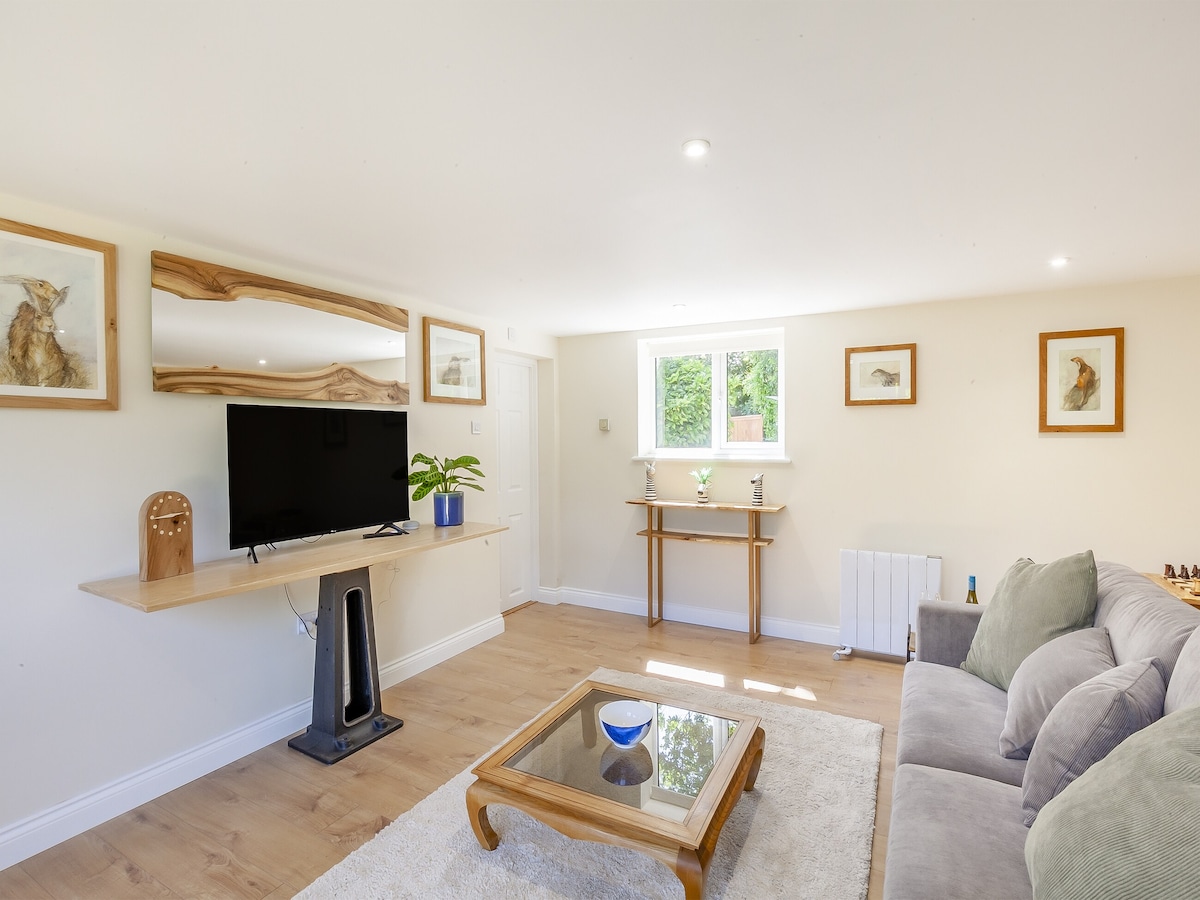 A spacious living room features a smart TV mounted on a wooden wall shelf, complemented by framed animal artwork. A small console table is placed beneath a window, allowing natural light to illuminate the light-colored walls and wooden flooring.